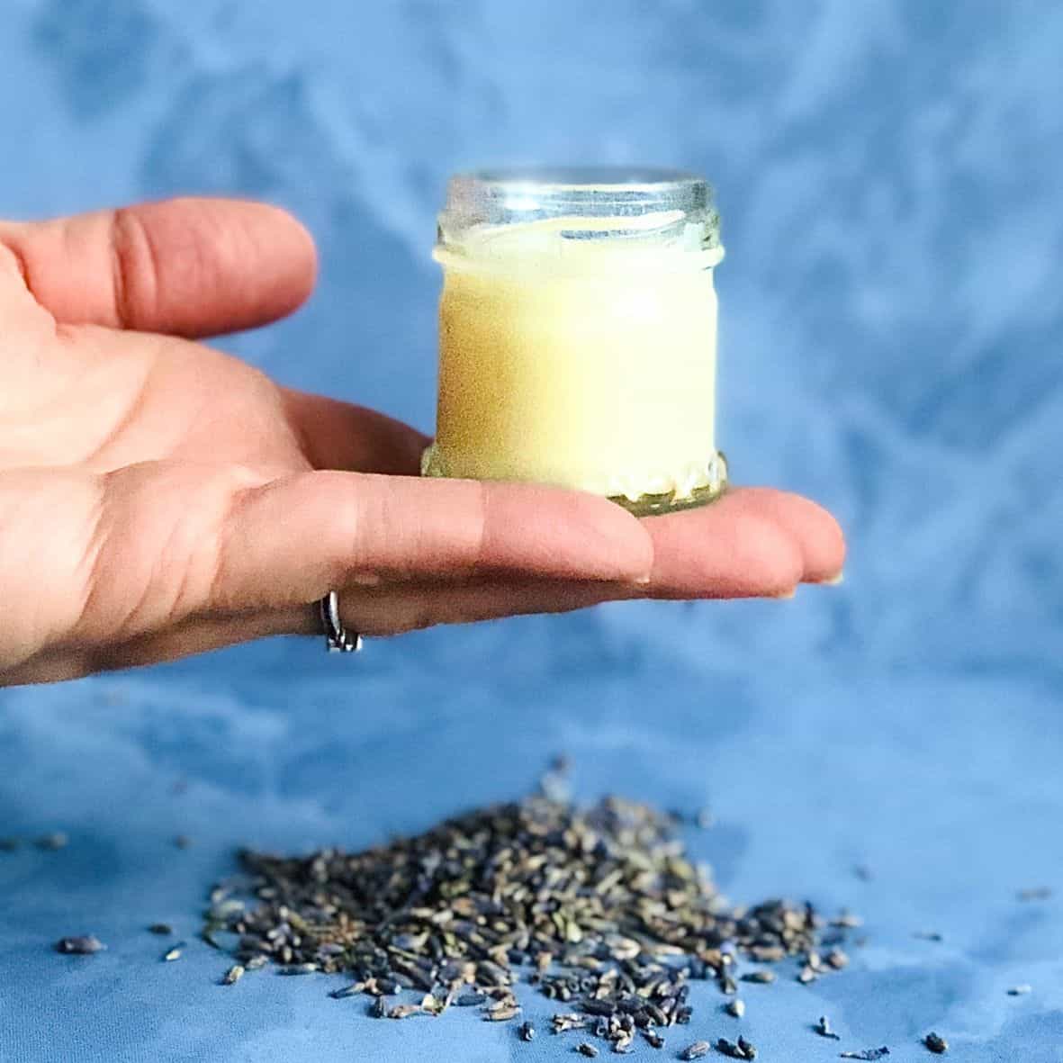A woman holding a homemade lip balm jar on her palm against blue tiles.