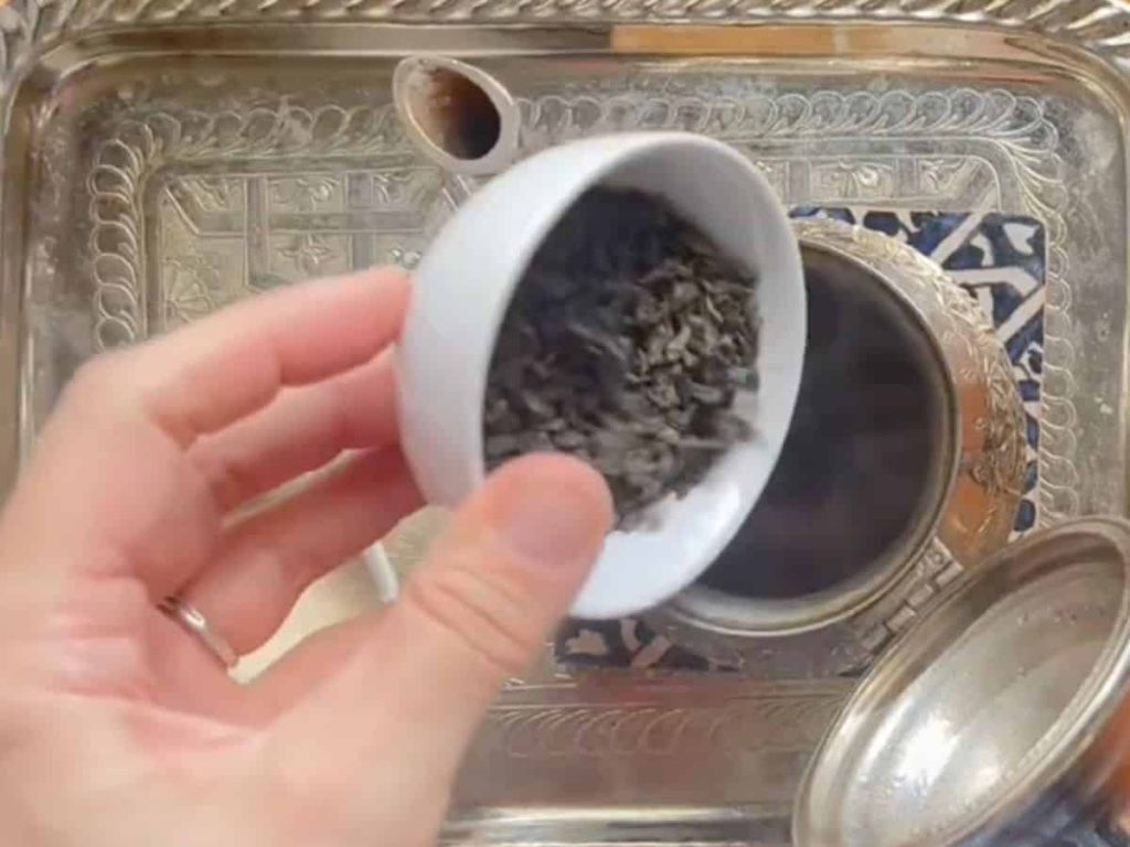 A woman is pouring loose-leaf green tea into the brass kettle.