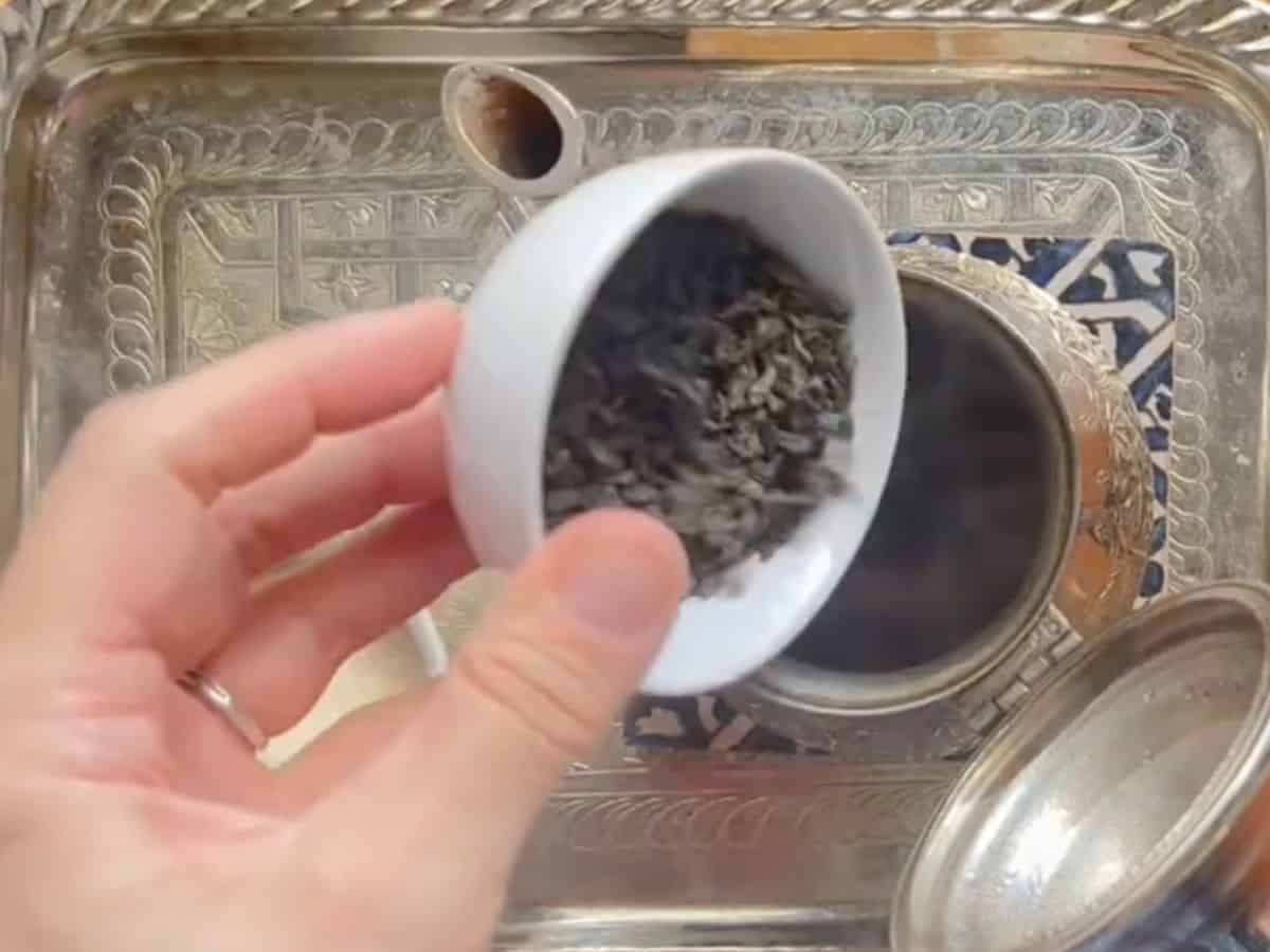 A woman is pouring loose-leaf green tea into the brass kettle.