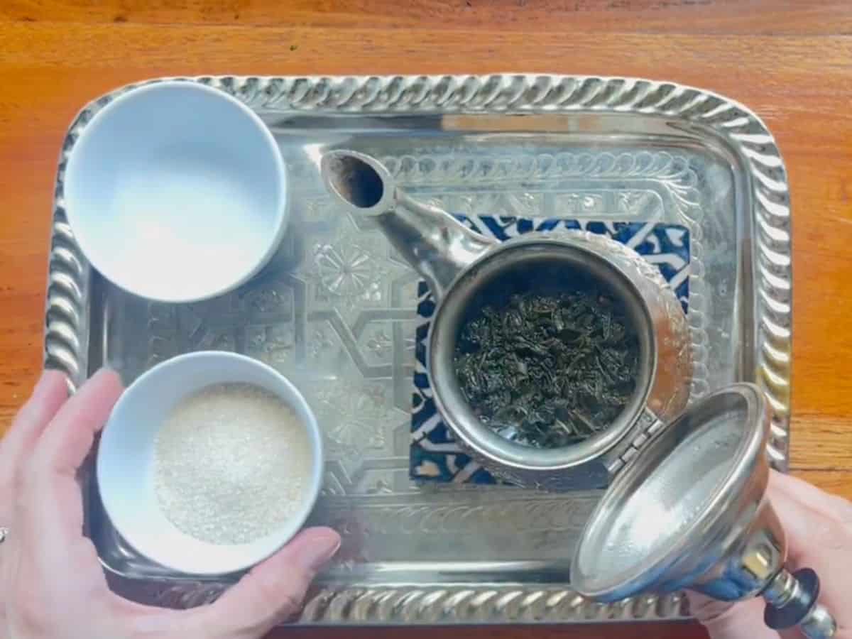 A woman is adding granulated sugar into the Moroccan tea kettle with water and green tea.