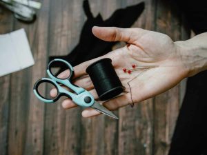Open hand holding tiny scissors, yarn, and pins on the palm to repair family clothes to save money.