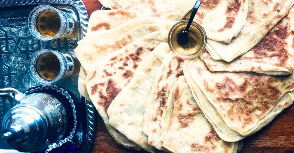 Crispy square Moroccan bread msemen arranged in a circle with the honey jar in the middle. On the side a tray with Moroccan mint tea pot and two glasses.