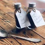 Two glass bottles filled with Hot Chocolate Mix with Hot Chocolate Tags and a silver spoon lying on the side on a wooden surface.