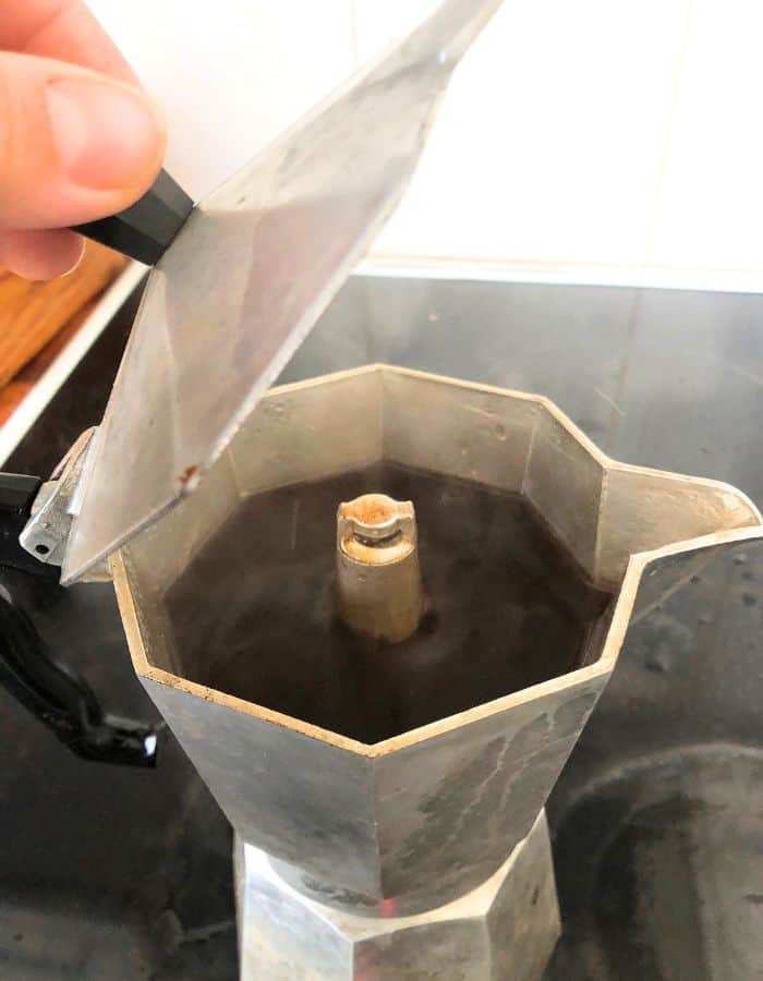 A woman opening the lid of an Italian coffee maker to see if the coffee is ready.