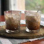 Two glasses filled with ice cubes, iced coffee and almond milk and spoons sticking out of them on a wooden tray.