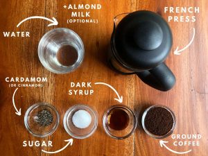 French Press coffee maker, glass bowls with ground coffee, cardamom, sugar, and dark syrup, and a glass of hot water placed on the table.