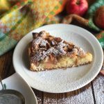 Piece of Finnish apple pie served on a white plate, the rest of the pie is on the background with a fall. colored tea towel and red apples.