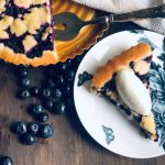 Blueberry pie on a cake tin with a slice of the pie on a plate with vanilla ice cream quenelle.