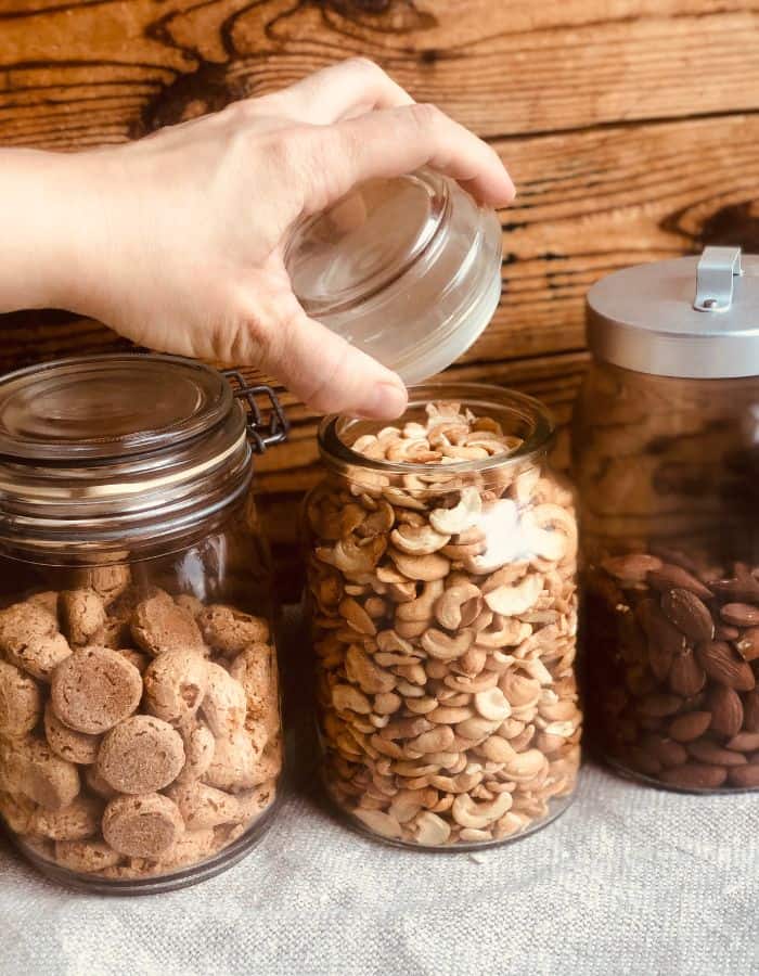 A woman lifting the lid of a cashew jar in a line of glass jars with almonds and biscuits.