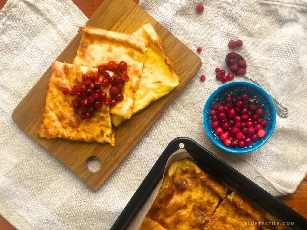 Swedish pancakes cut in squares on a wooden board with some lingonberries on the top and in a small blue bowl.