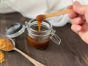 Homemade natural brown sugar scrub paste in a glass jar and a woman is lifting with a wooden spoon some.