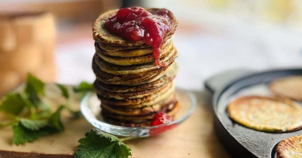 Nettle pancakes piled on top of each other and raspberry jam dripping from the top, on the side a cast iron Swedish pancake pan with pancakes frying.