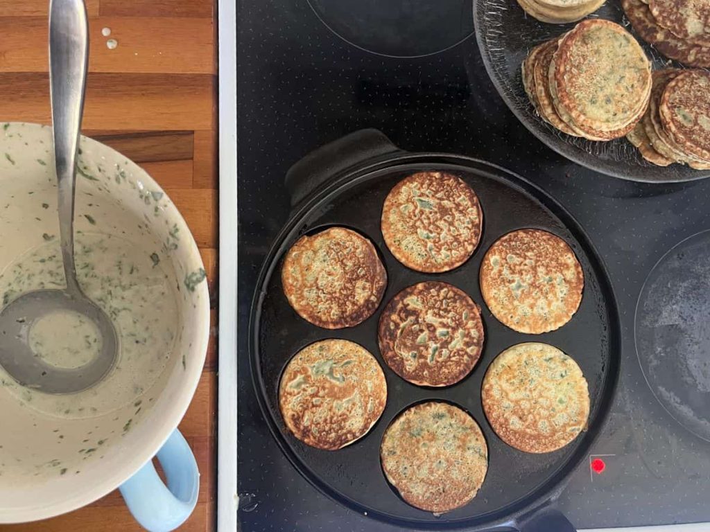 Cooking flow when frying nettle pancakes: batter bowl on the left, then panckaes pan on the stove frying and a plate of pancakes on the right with ready fried nettle pancakes.