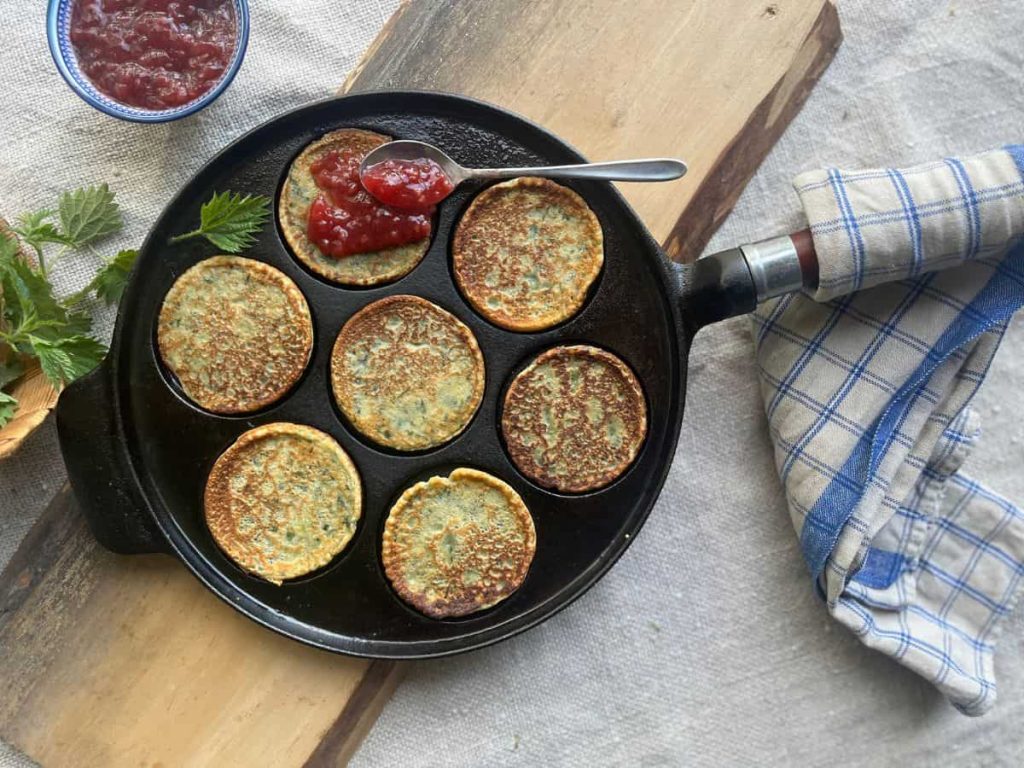 Nettle pancakes in a cast iron Swedish pancake pan on a wooden plank with a tea towel over the handle. A spoonful of raspberry jam is lying carelessly on the top of one pancake.