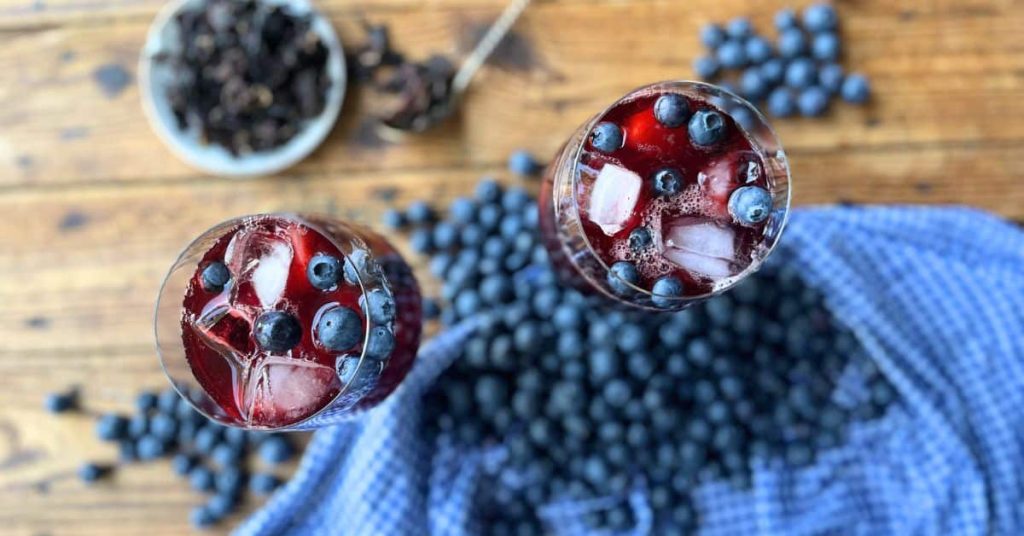 Two glasses filled with blueberries, ice cubes, and blueberry hibiscus iced tea in the middle, surrounded by hibiscus petals and blueberries on a wooden surface.