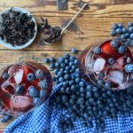 Two glasses of blueberry hibiscus iced tea in the middle with some blueberries, hibiscus petals on the wooden surface with a blue square pattern table cloth.