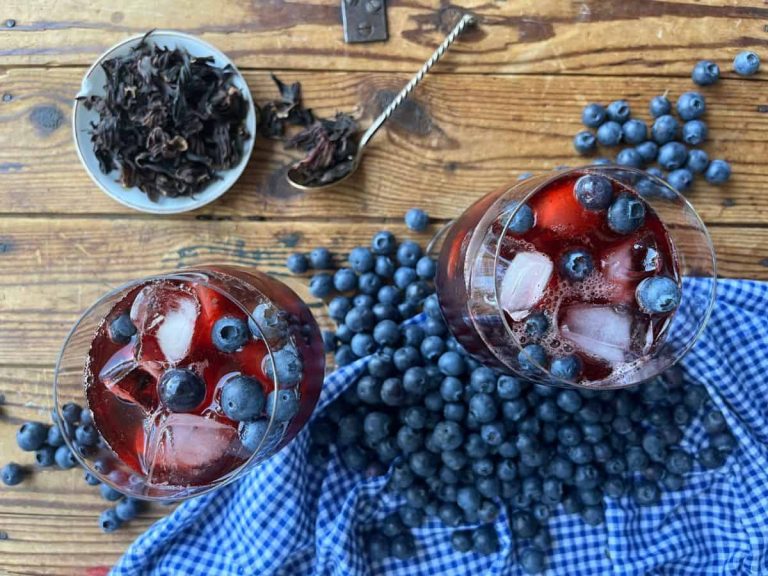 Two glasses of blueberry hibiscus iced tea in the middle with some blueberries, hibiscus petals on the wooden surface with a blue square pattern table cloth.