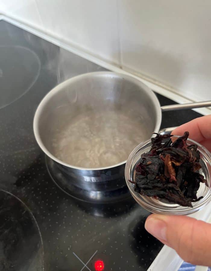 Water is boiling in a saucepan. Fingers are holding a small glass bowl with hibiscus petals for hibiscus tea.