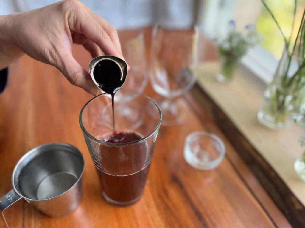 A woman is pouring homemade blueberry syrup into the hibiscus tea to make iced tea. 
