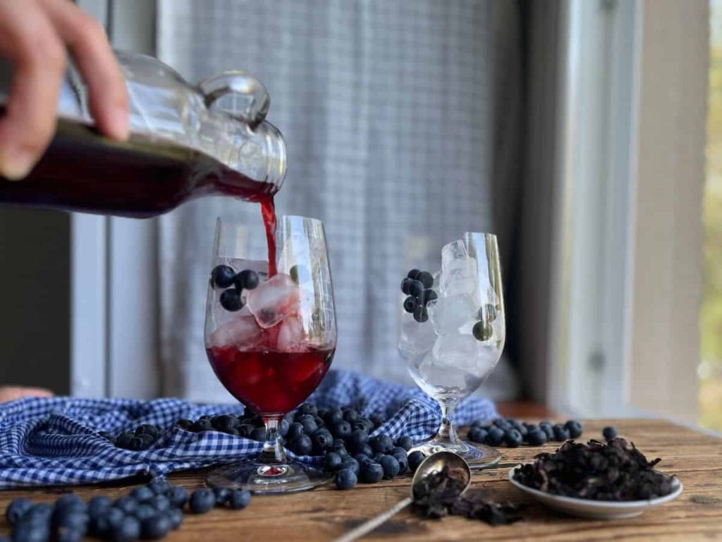 A woman is pouring the iced tea from a glass bottle into the glasses filled with ice and blueberries.