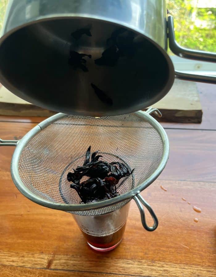 A strainer over a mixing glass to catch the hibiscus petals when woman is pouring the hibiscus tea from the saucepan.