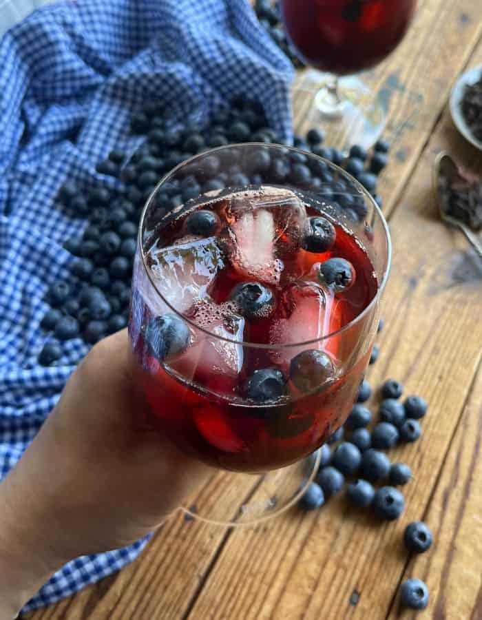 A woman holding a glass of blueberry iced tea with hibiscus.