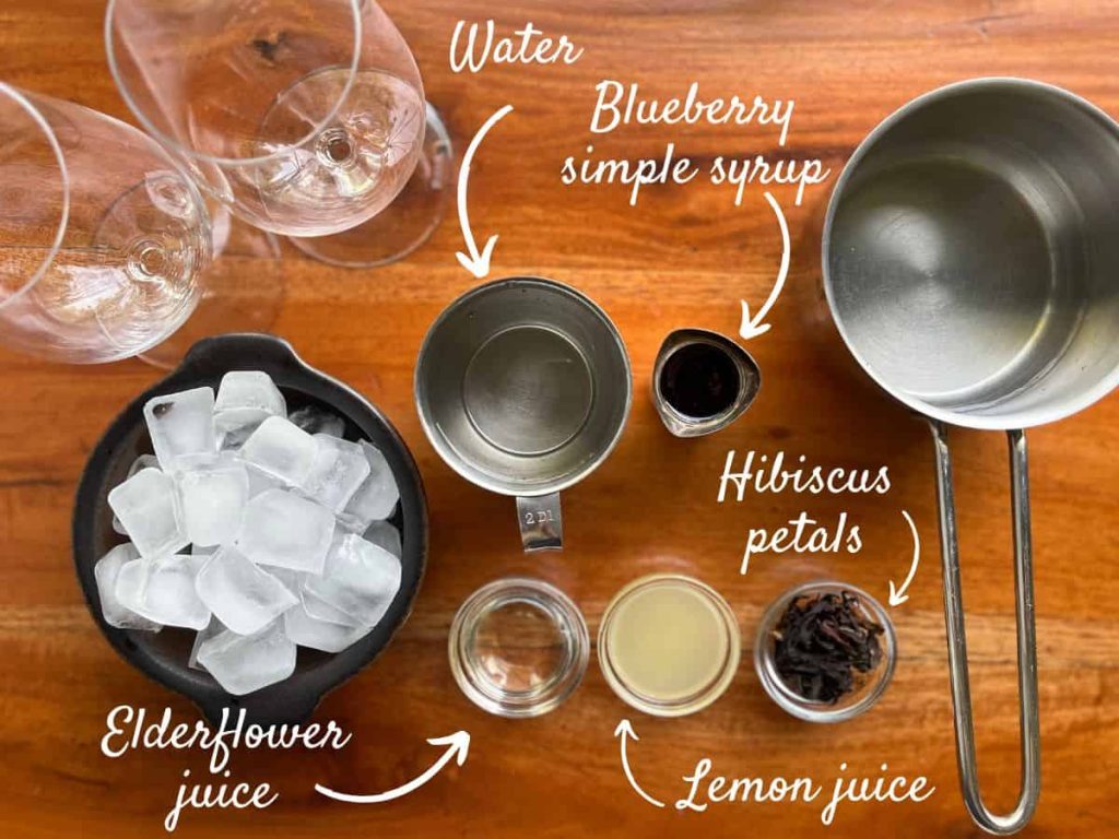 Ingredients for the homemade hibiscus blueberry iced tea: Water, ice cubes, blueberry syrup, hibiscus petals, lemon juice, and elderflower juice.
