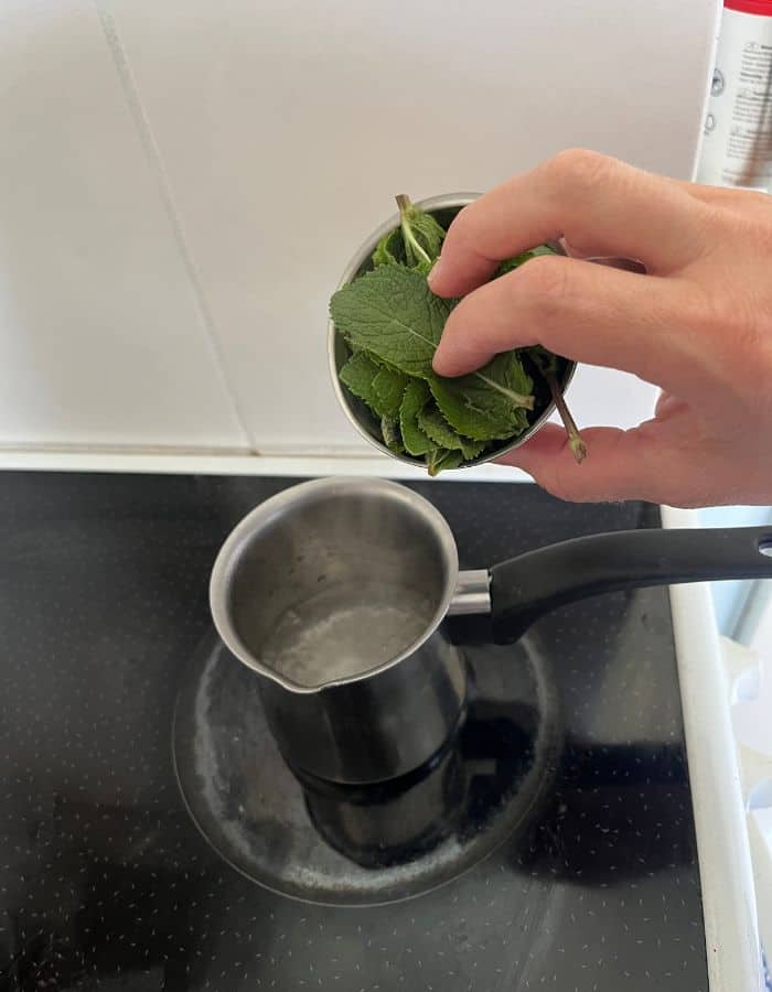 A woman is pouring mint leaves into a small saucepan on the stove.