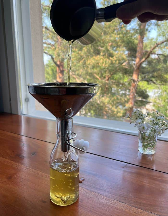A woman is pouring the finished mint and peppermint simple syrup through the funnel into a glass bottle.