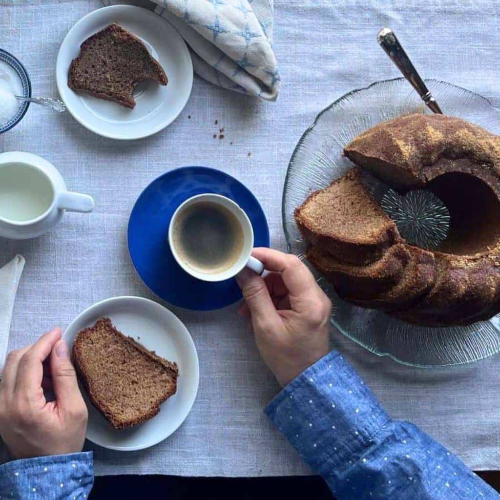 Spiced coffee cake on a serving platter and a sliced cut on a plate in front of a woman who is holding a coffee cup on a blue saucer.
