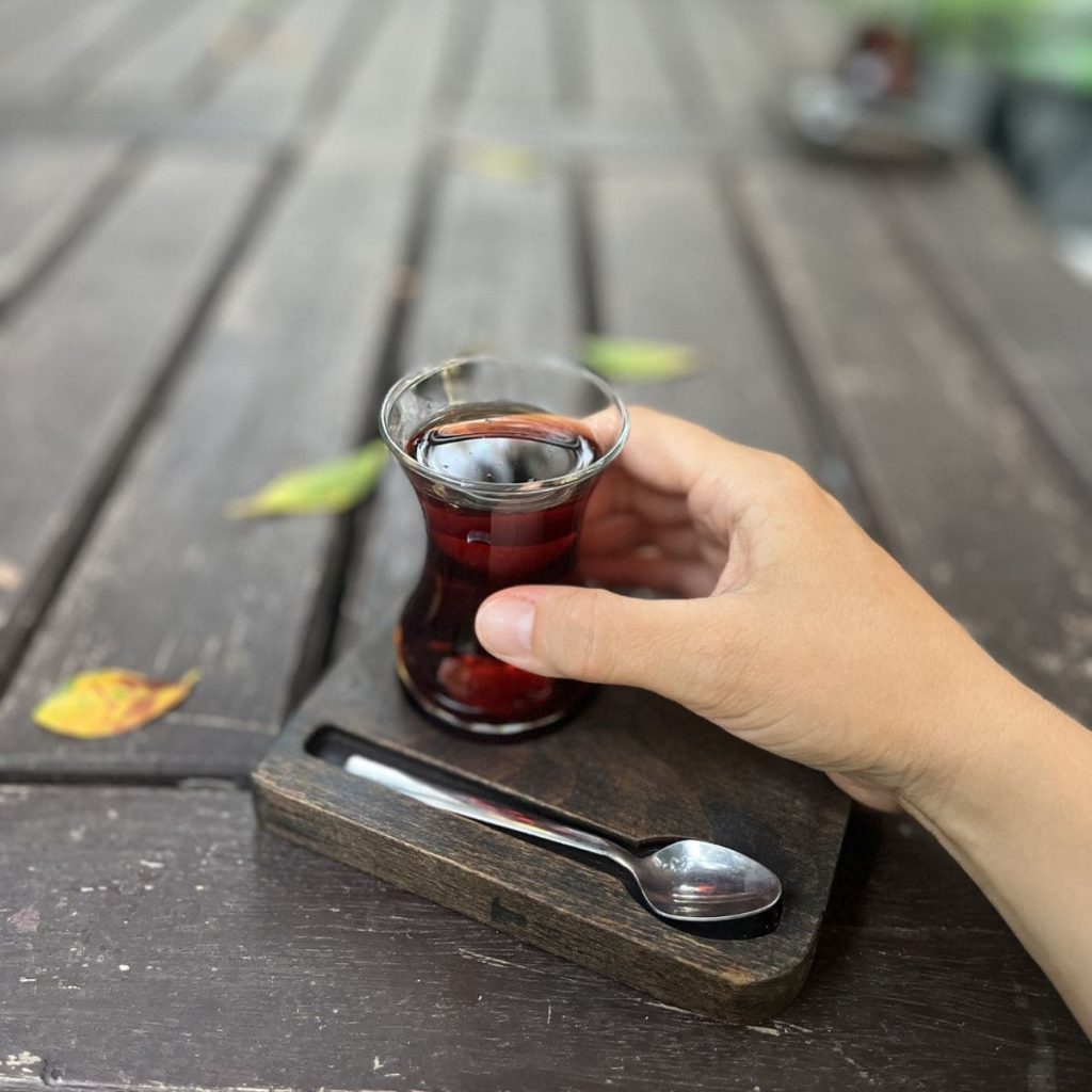 A woman outside holding in her hand a black tea glass with a spoon hand relaxed on a patio table.