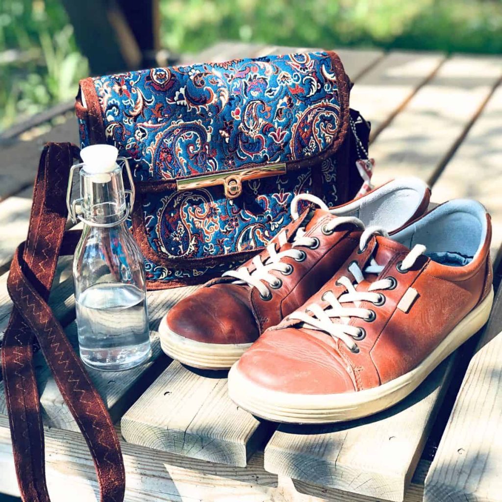 Handbag, glassbottle filled with water and leather shoes on a sunny wooden floor outside.