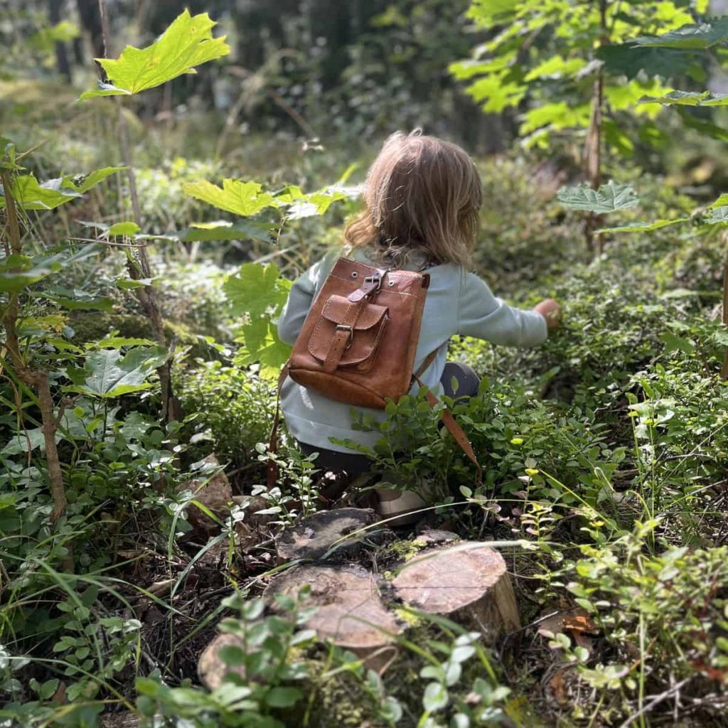 A young girl with a leather backpack knelt down on a hike in the forest. 