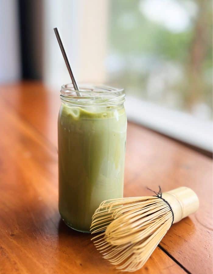 Iced vanilla matcha latte in a tall glass with a bamboo whisk, matcha chasen on the side.