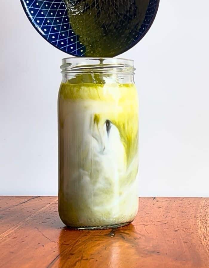 An iced vanilla matcha latte on the table and a woman is pouring the vanilla matcha mixture in to create an Instagrammable photo of the Iced Vanilla Matcha Latte.