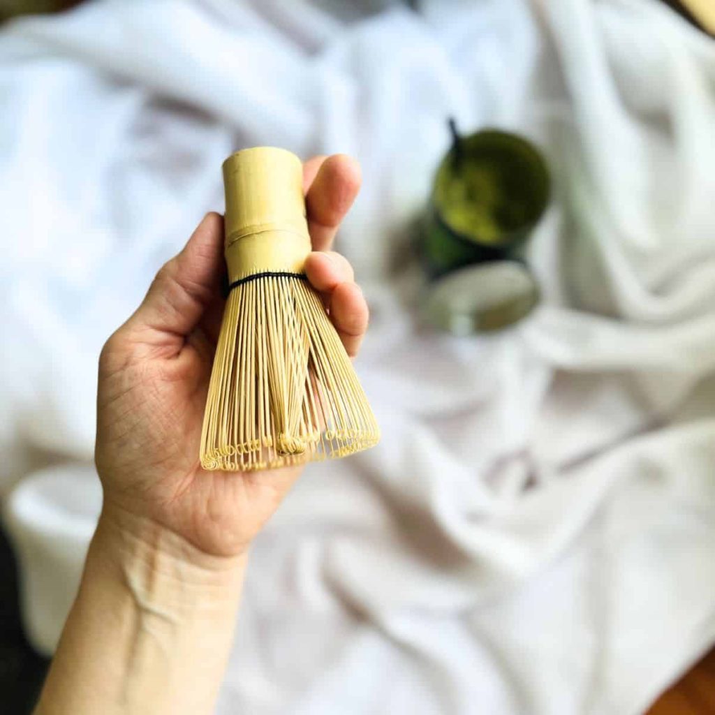 A woman holding a matcha chasen the bamboo whisk in her hand agains white linen and an open matcha tin in the background.