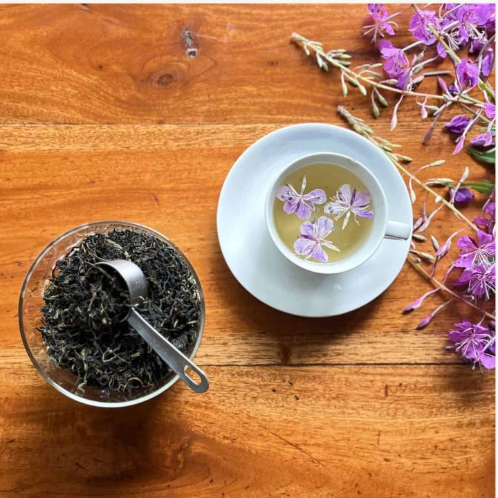A jar of homemade Ivan Chai loose-leaf herbal tea with a steeped cup of the same fermented fireweed tea with violet colored fireweed flowers as garnish and on the side.
