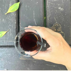 A woman is holding a black tea glass in her hand against a wooden table with a few fallen leaves.