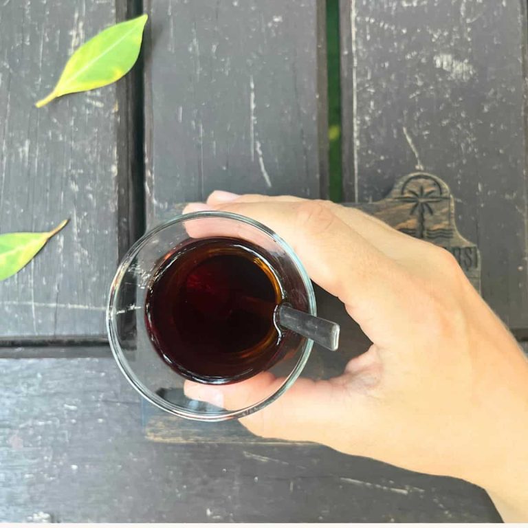 A woman is holding a black tea glass in her hand against a wooden table with a few fallen leaves.