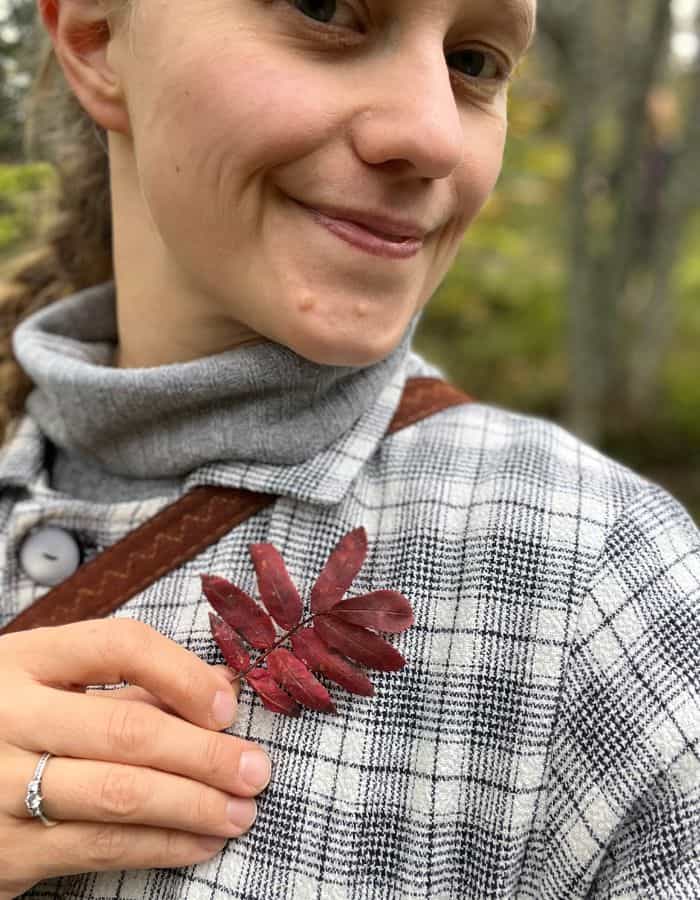 A woman smiling happily and holding an autumn leaf against her light colored jacket.