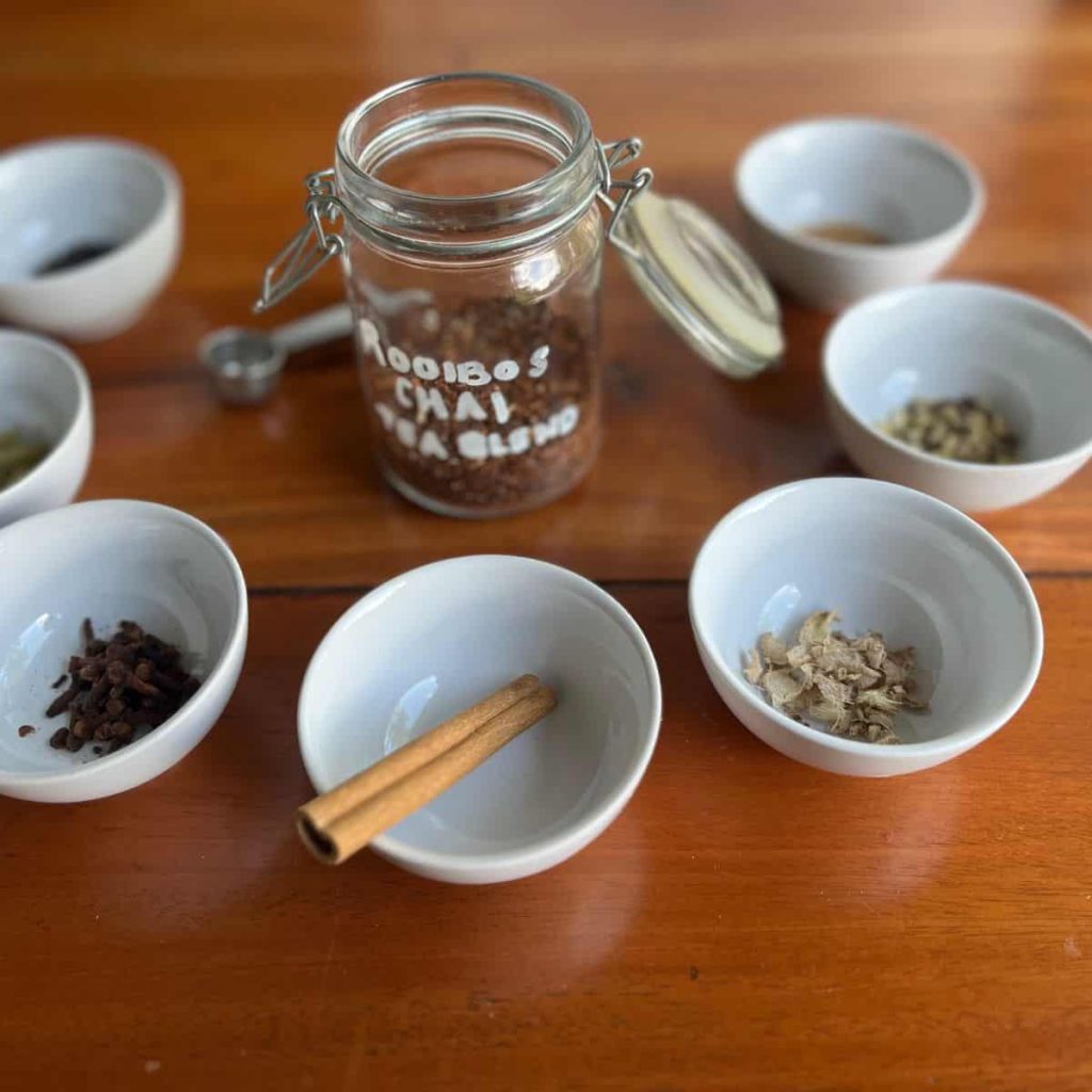 Rooibos chai tea blend in making: Spiced in small bowls around the glass jar filled with rooibos tea leaves.