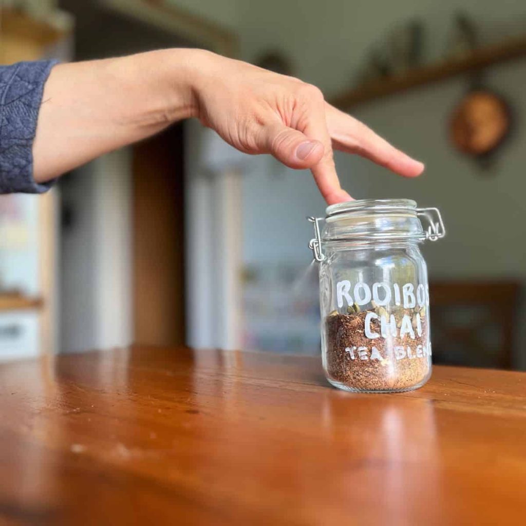 A woman holding a finger on the top of a jar of homemade rooibos chai tea blend.