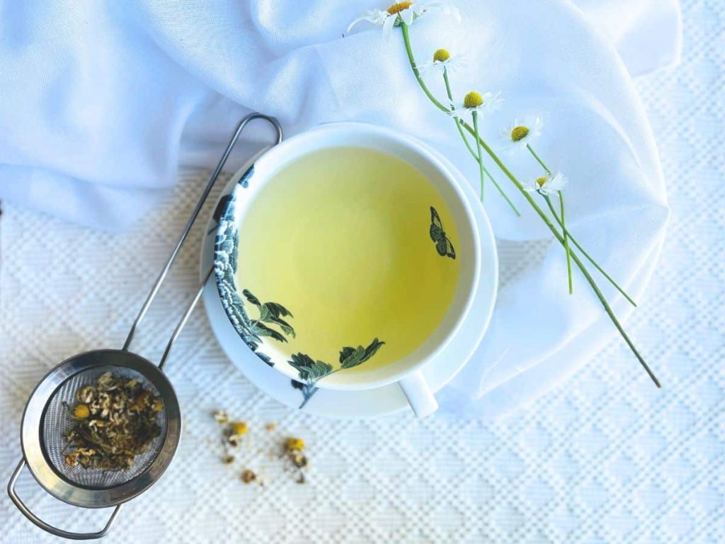 A teacup filled with chamomile herbal tea and dried chamomile buds in teastrainer on the side against white background.